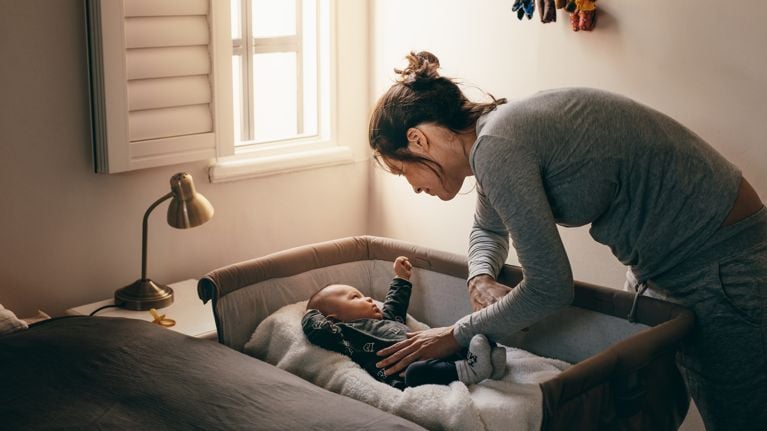 Mother putting her baby to sleep on a bedside baby crib. Woman bending forward over a crib to check her sleeping baby.