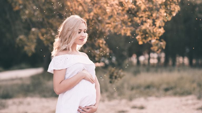 Smiling pregnant woman 20-24 year old wearing white stylish dress standing outdoors over nature background close up. Motherhood. Maternity.
