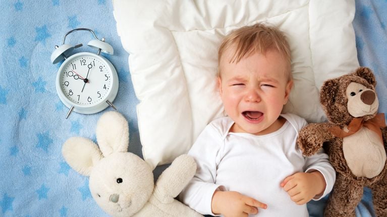 One year old baby lying in bed with alarm clock and crying
