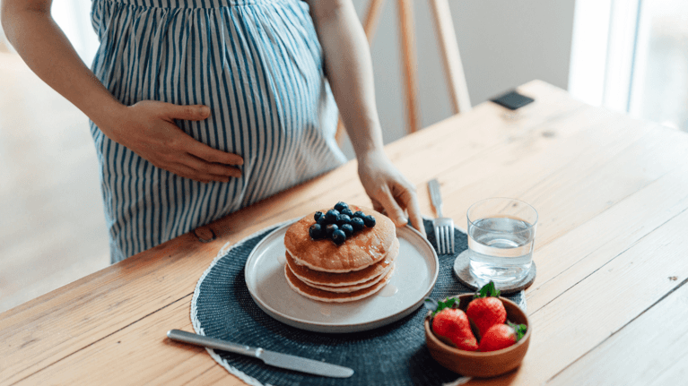 pregnant woman standing at the kitchen counter with a plate of pancakes and fruit in front of her